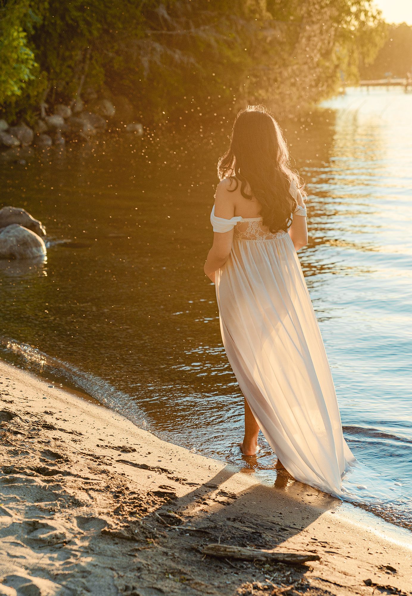 Pregnant woman in flowing white dress walking along sandy beach shore at golden sunset.