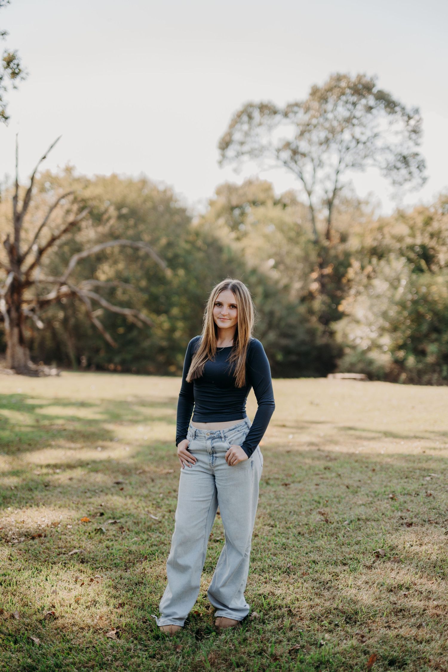 Young woman standing outdoors near trees, smiling, wearing a long-sleeved black top and light-colored jeans.