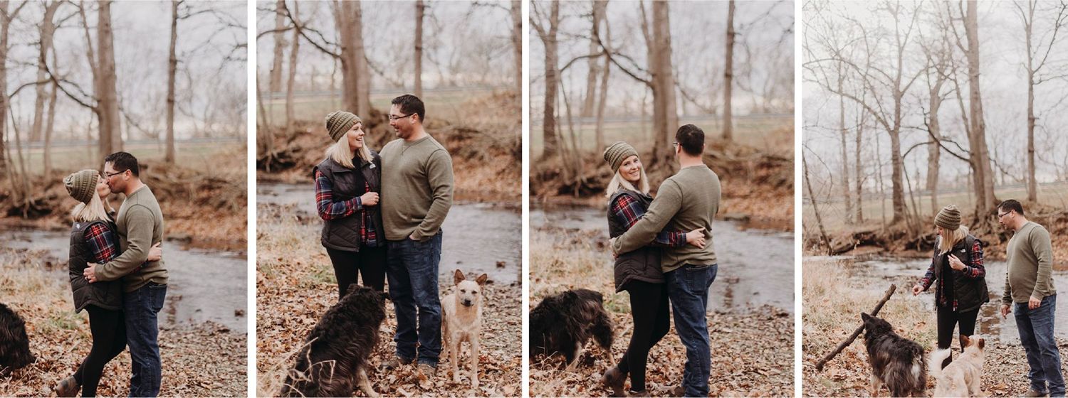A man and a woman standing near a creek with two dogs, surrounded by leafless trees on a cloudy day.