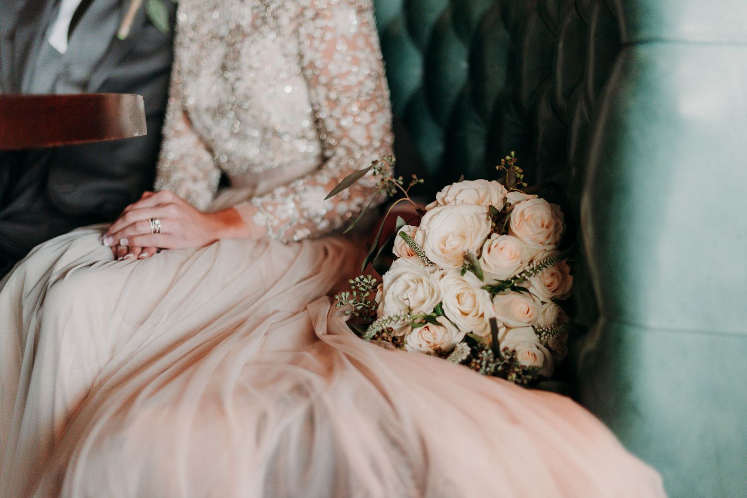 A man and a woman sit in a green booth on their wedding day with off white beaded dress and matching florals.