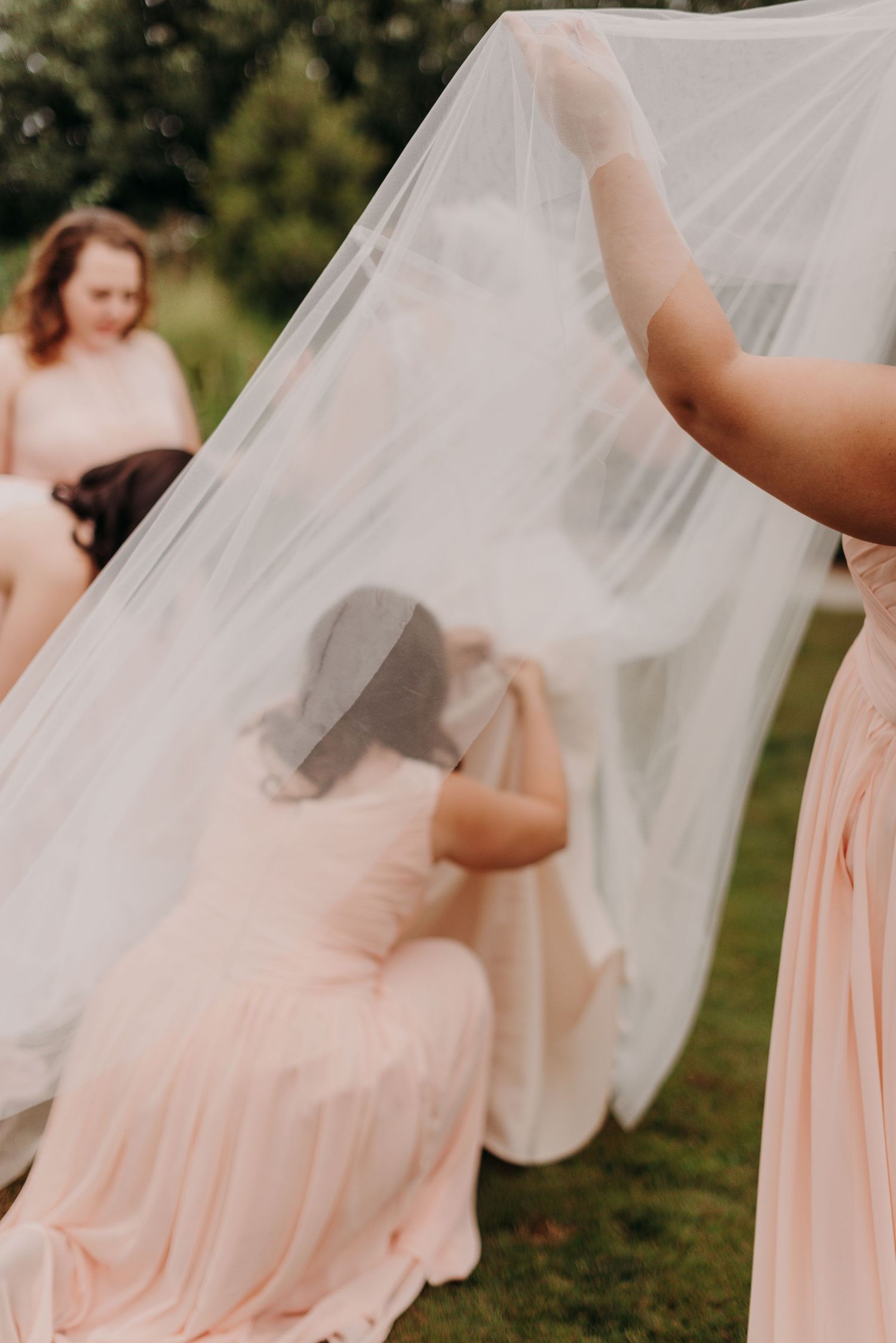 A bridesmaid is under the brides veil adjusting the brides dress on the day of her wedding.
