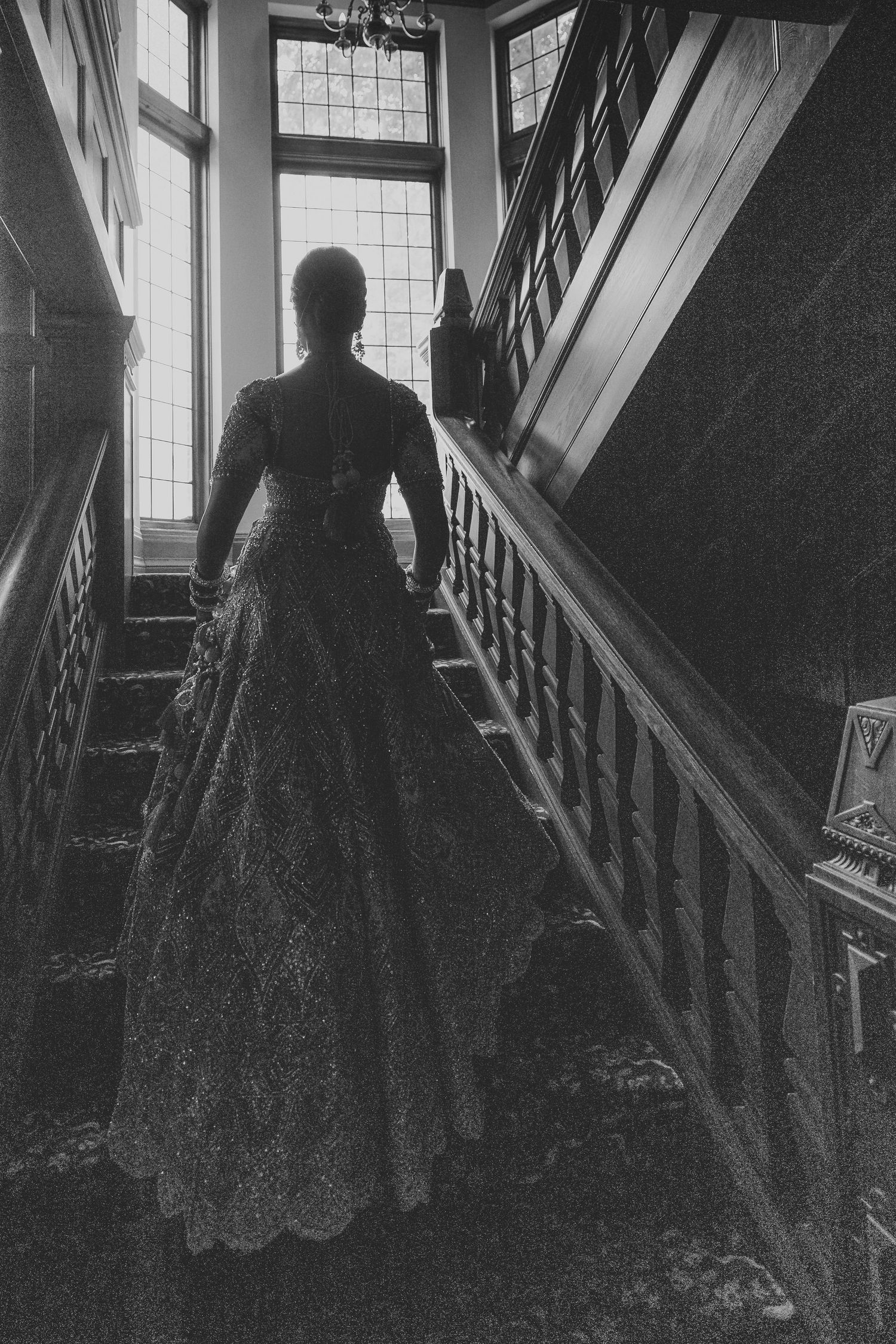 Black and white silhouette of bride in lace gown ascending grand staircase by window.