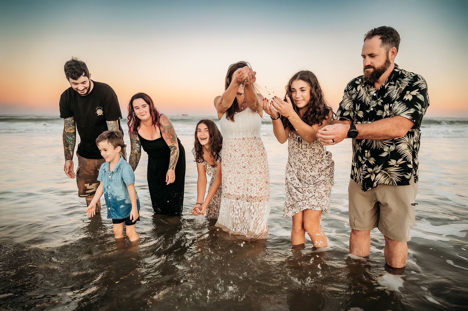 Family group photo at sunset on the beach, standing in shallow ocean waves, smiling and laughing together.
