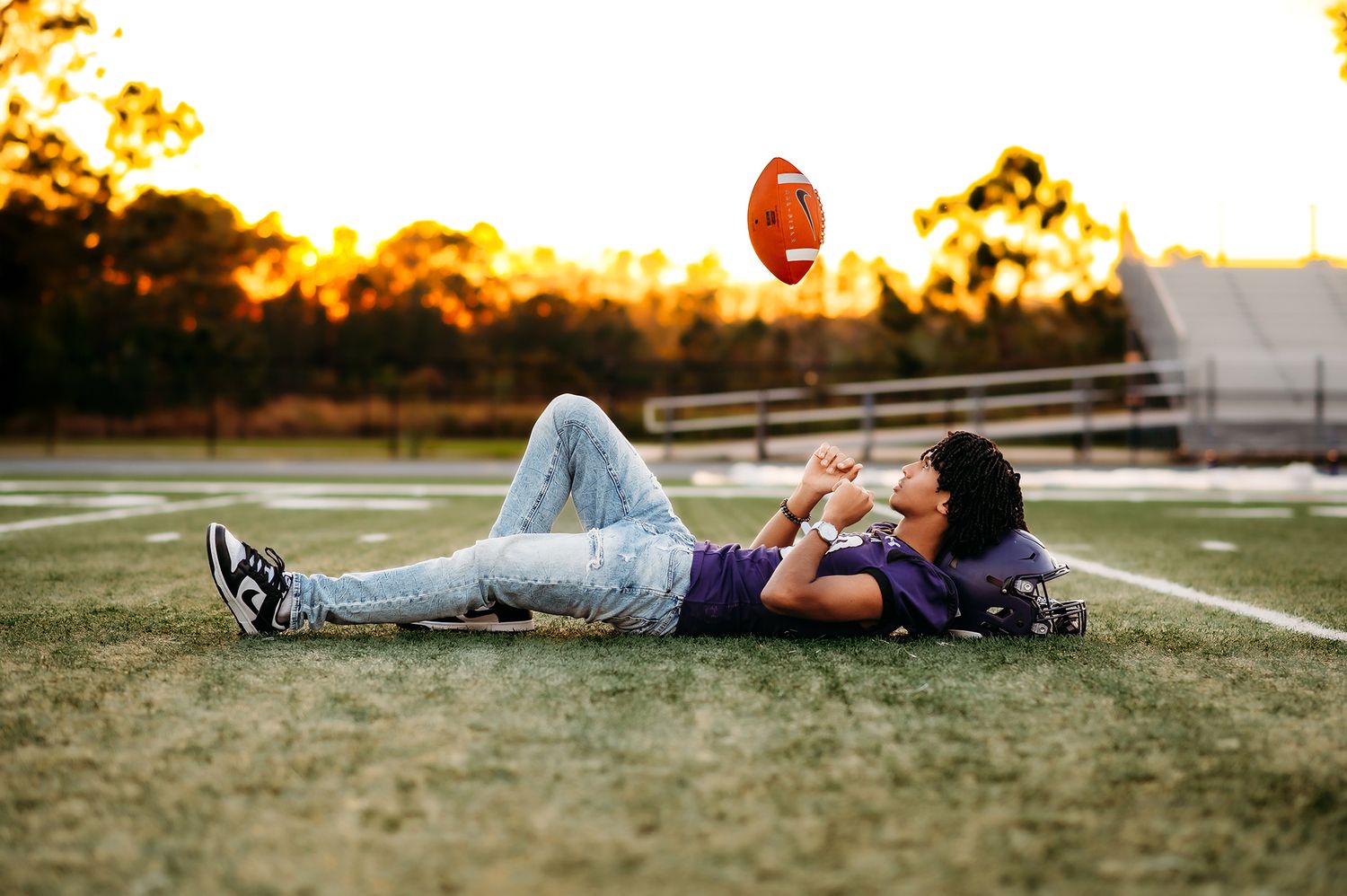 Teen boy lying on football field tossing a football in the air at golden hour sunset.