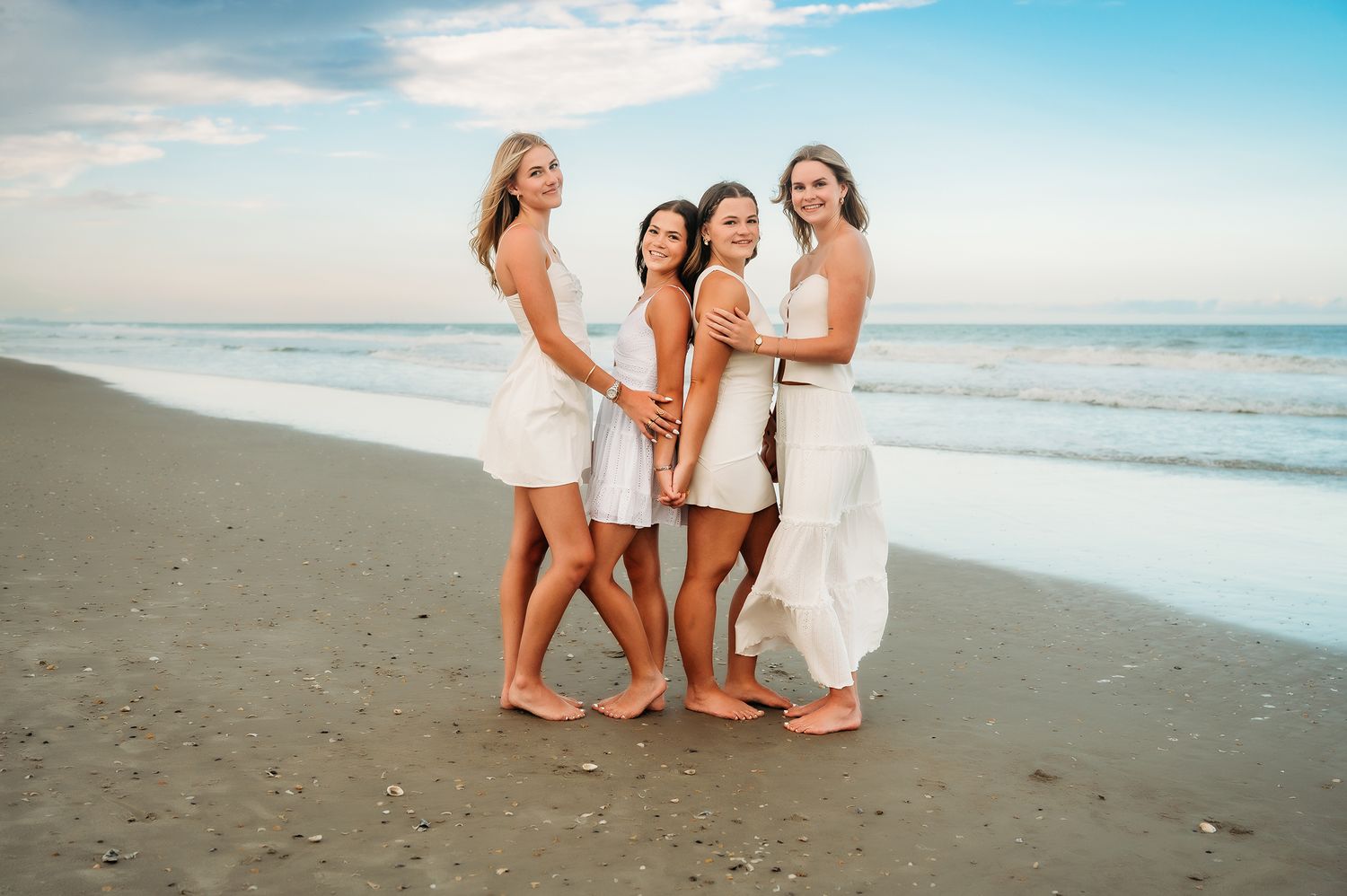 Four women in white outfits smiling and embracing on a sandy beach at sunset.