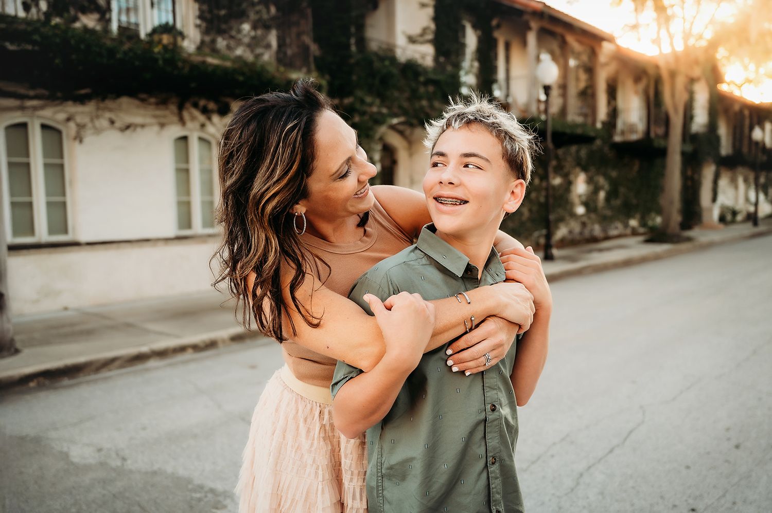 A woman and teenage boy share a joyful embrace on a sunlit street, smiling warmly in casual outdoor attire.