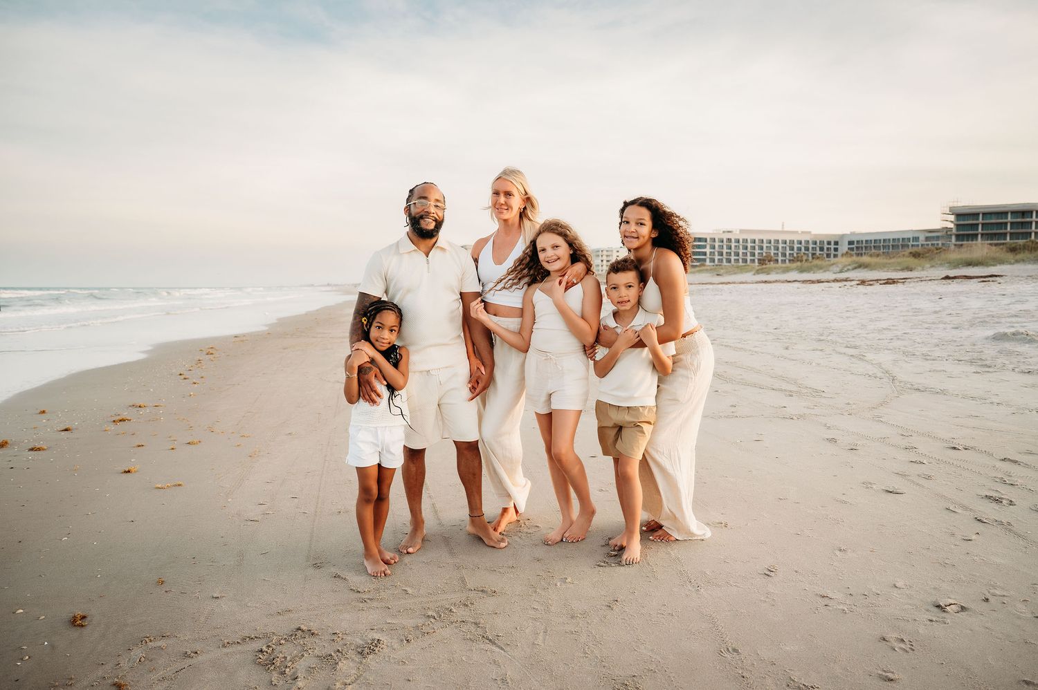 Happy family of six posing together on a sandy beach, wearing coordinated white outfits with ocean waves in the background.