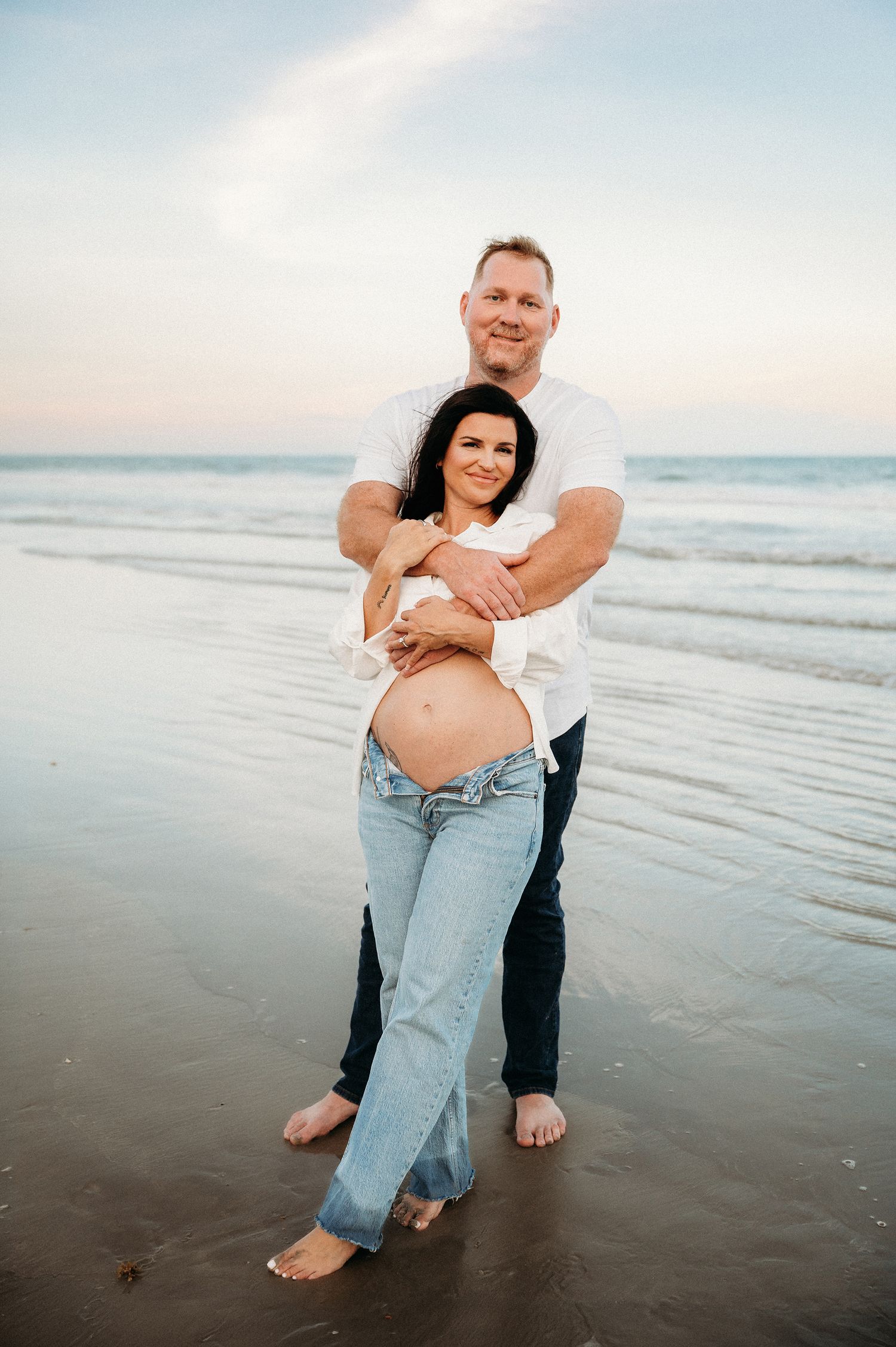 Couple sharing a tender beach embrace, man hugging pregnant woman from behind at sunset shoreline.