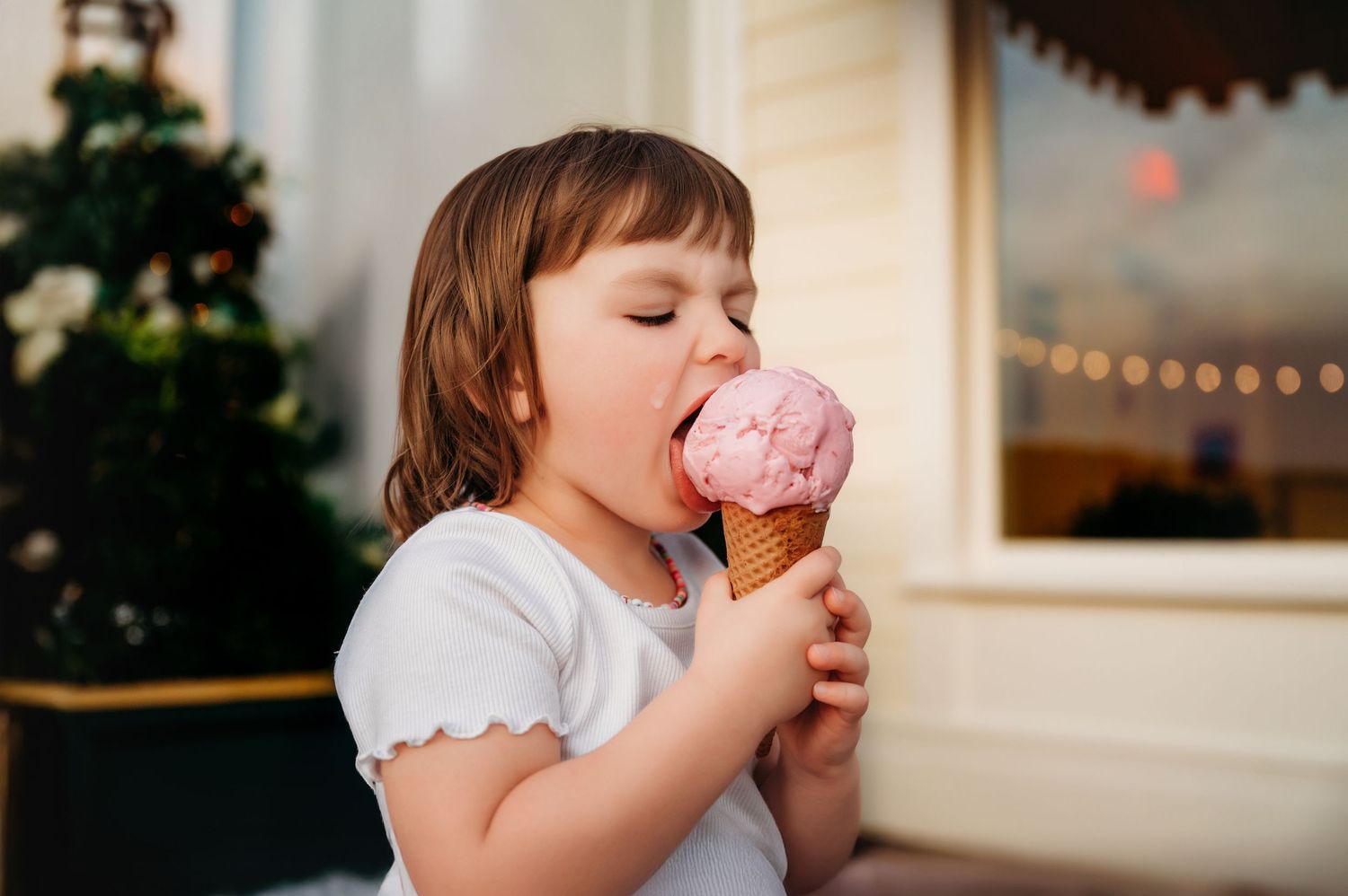 Adorable young girl with eyes closed, licking a large pink strawberry ice cream cone outdoors.