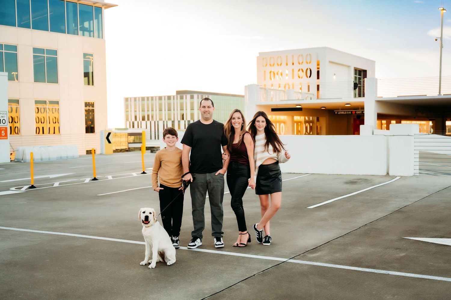 Family of four with a white dog posing in a parking garage in Orlando Florida at sunset near modern buildings.