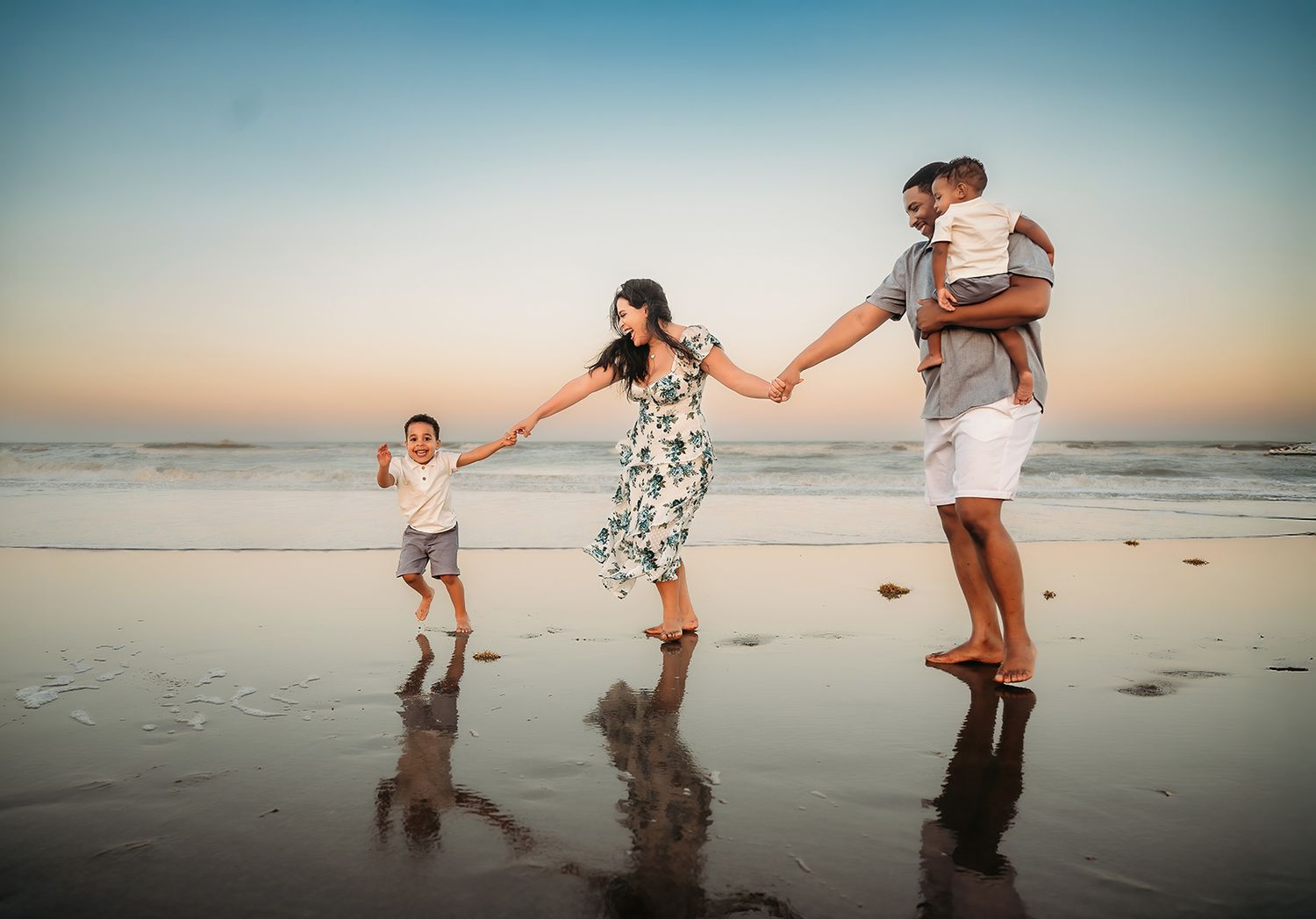 Family of four enjoying a sunset beach walk, parents holding hands with children on wet sand.