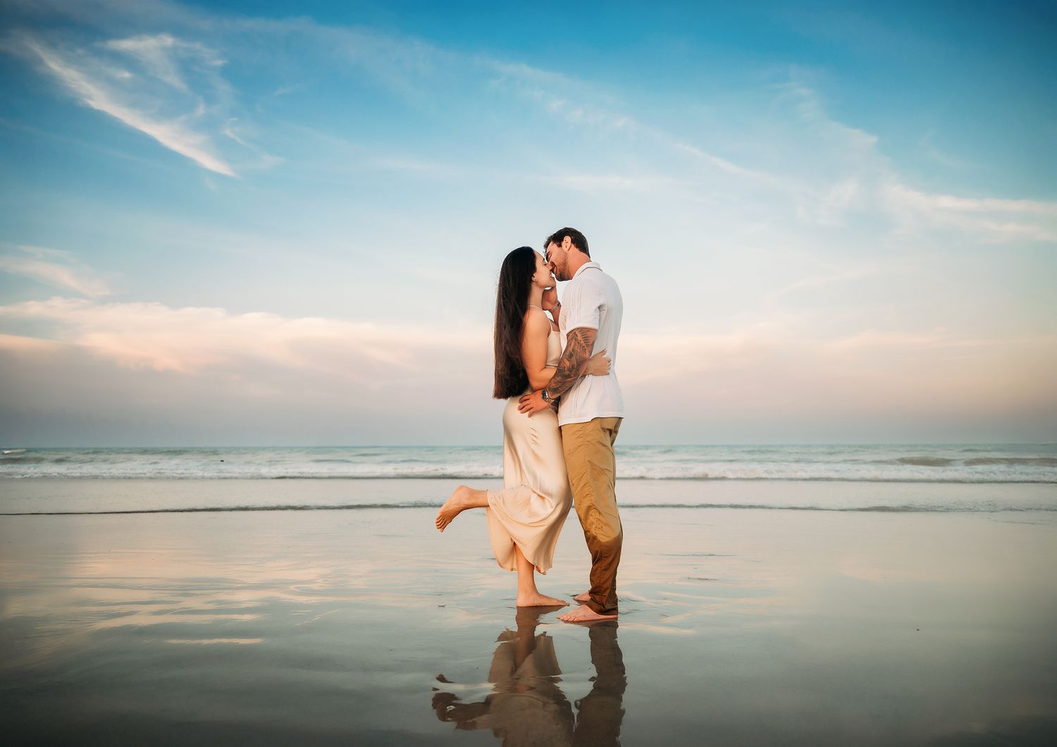 Couple sharing a romantic kiss on a reflective beach at sunset with ocean waves and pastel sky behind them.