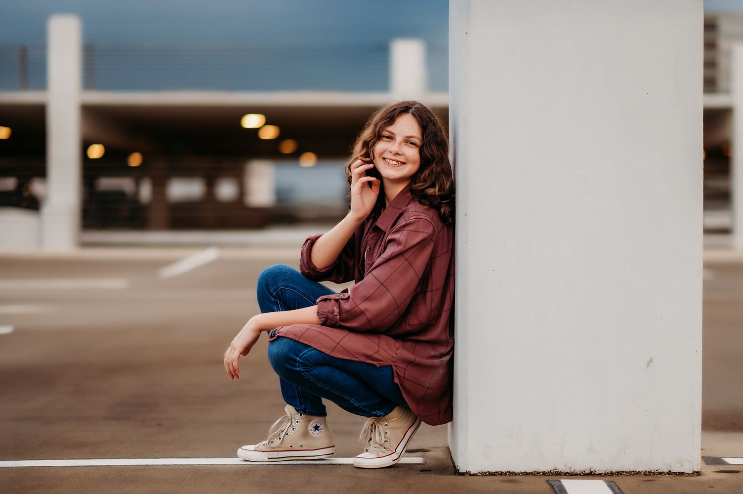 Smiling young woman sitting casually on a parking garage floor leaning against a concrete pillar.
