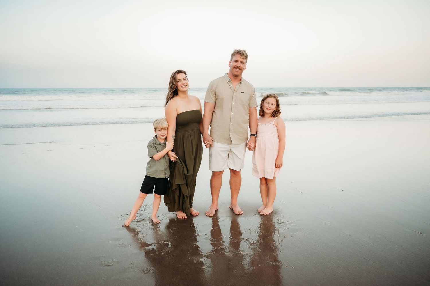 Family of four posing on a reflective beach at sunset, parents and two children smiling in coordinated neutral outfits.