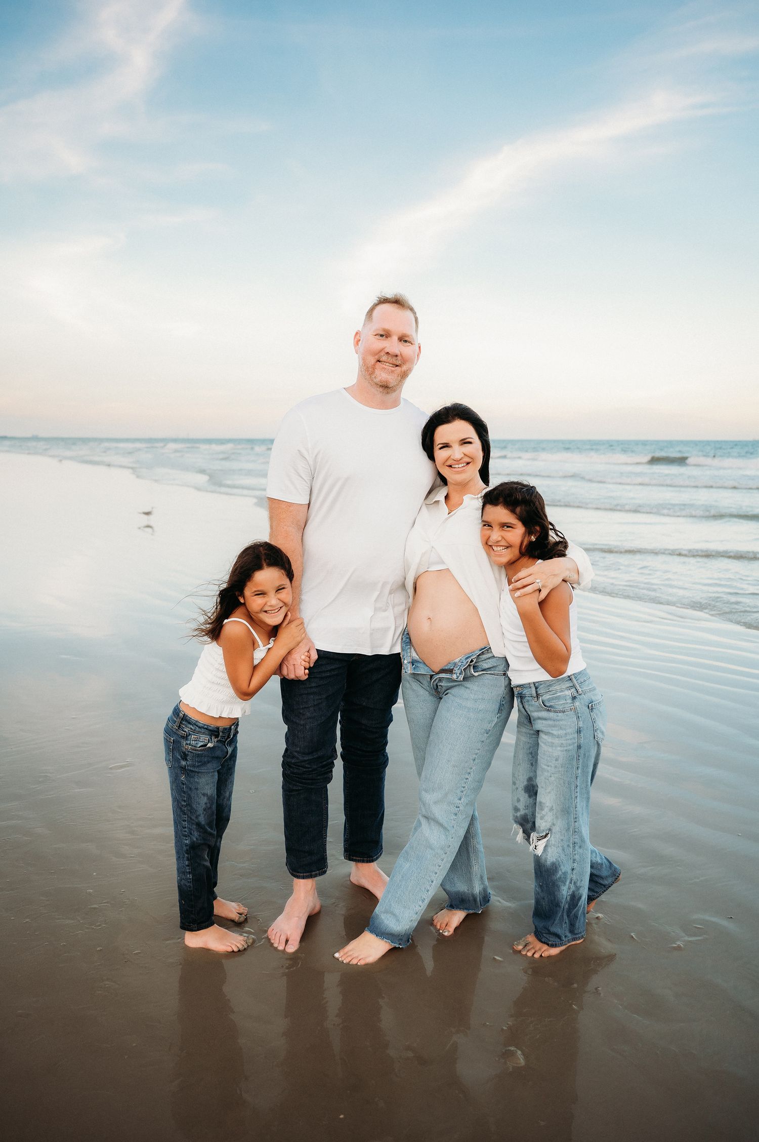 Pregnant woman with husband and two daughters posing barefoot on a beach at sunset in jeans and white tops.