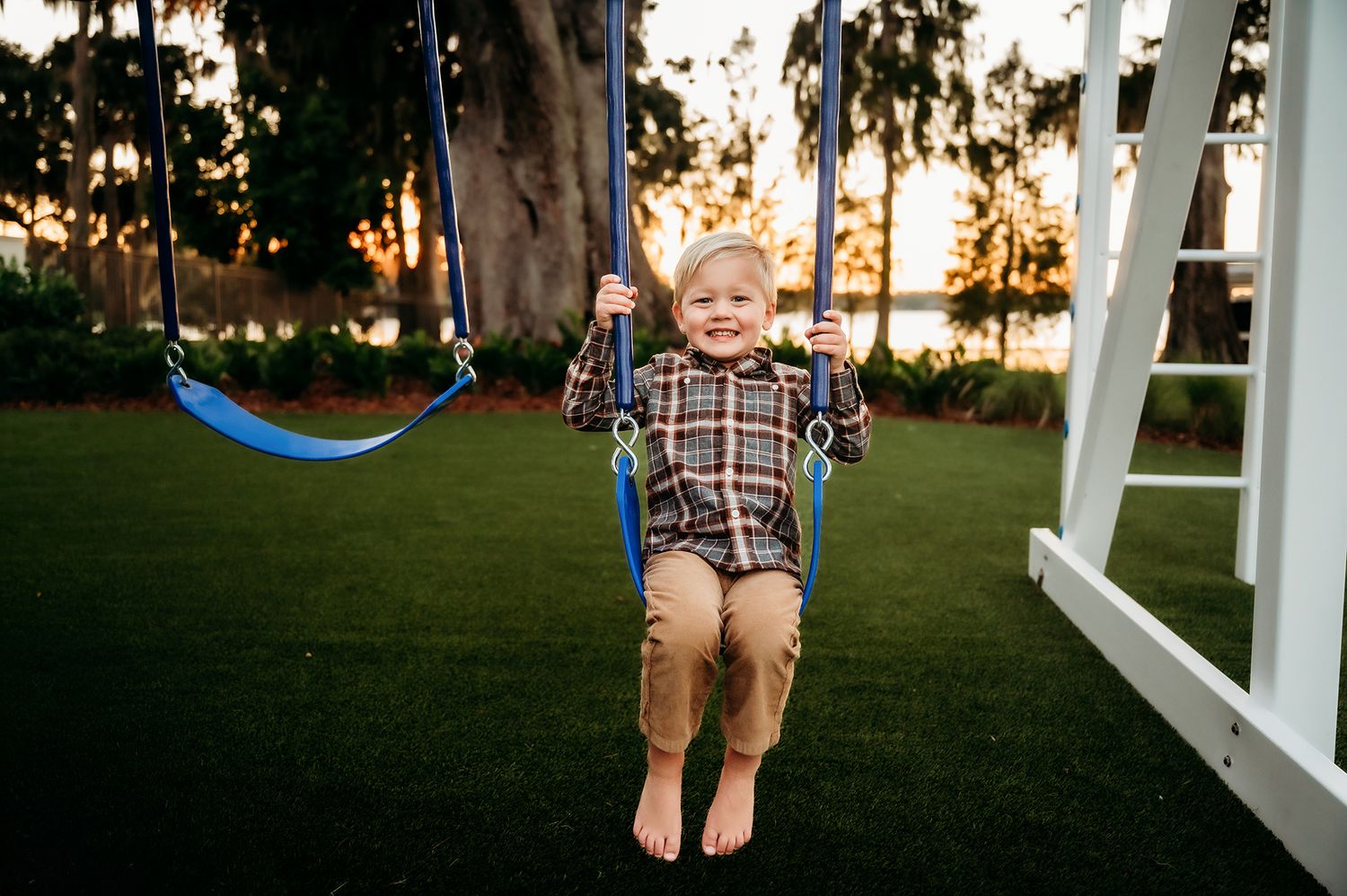 Happy toddler boy smiling on a blue swing set outdoors on green grass during golden hour.