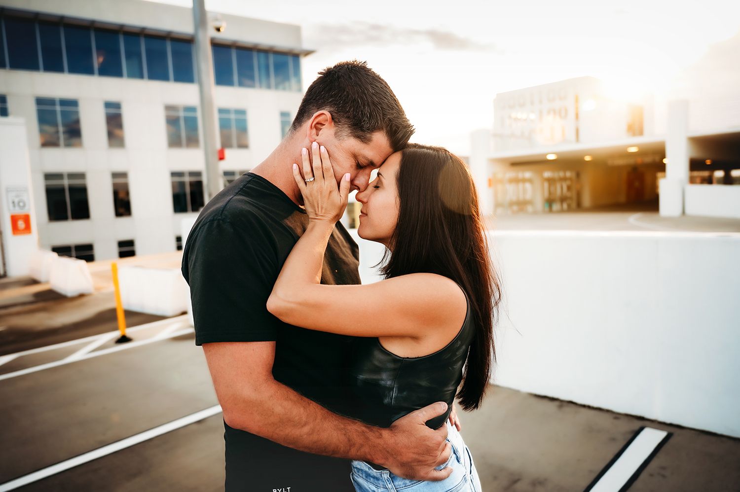 Couple sharing a romantic kiss outdoors in a parking lot with warm sunlight glowing behind them.