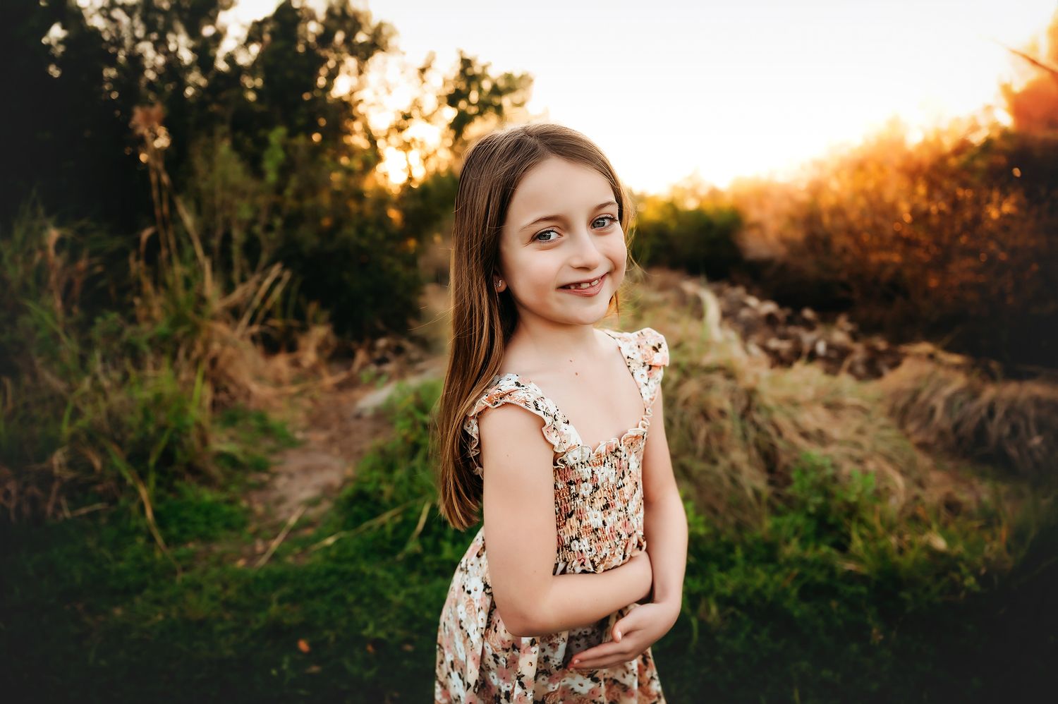 A young girl in a floral dress smiles outdoors at golden hour with lush greenery in the background.