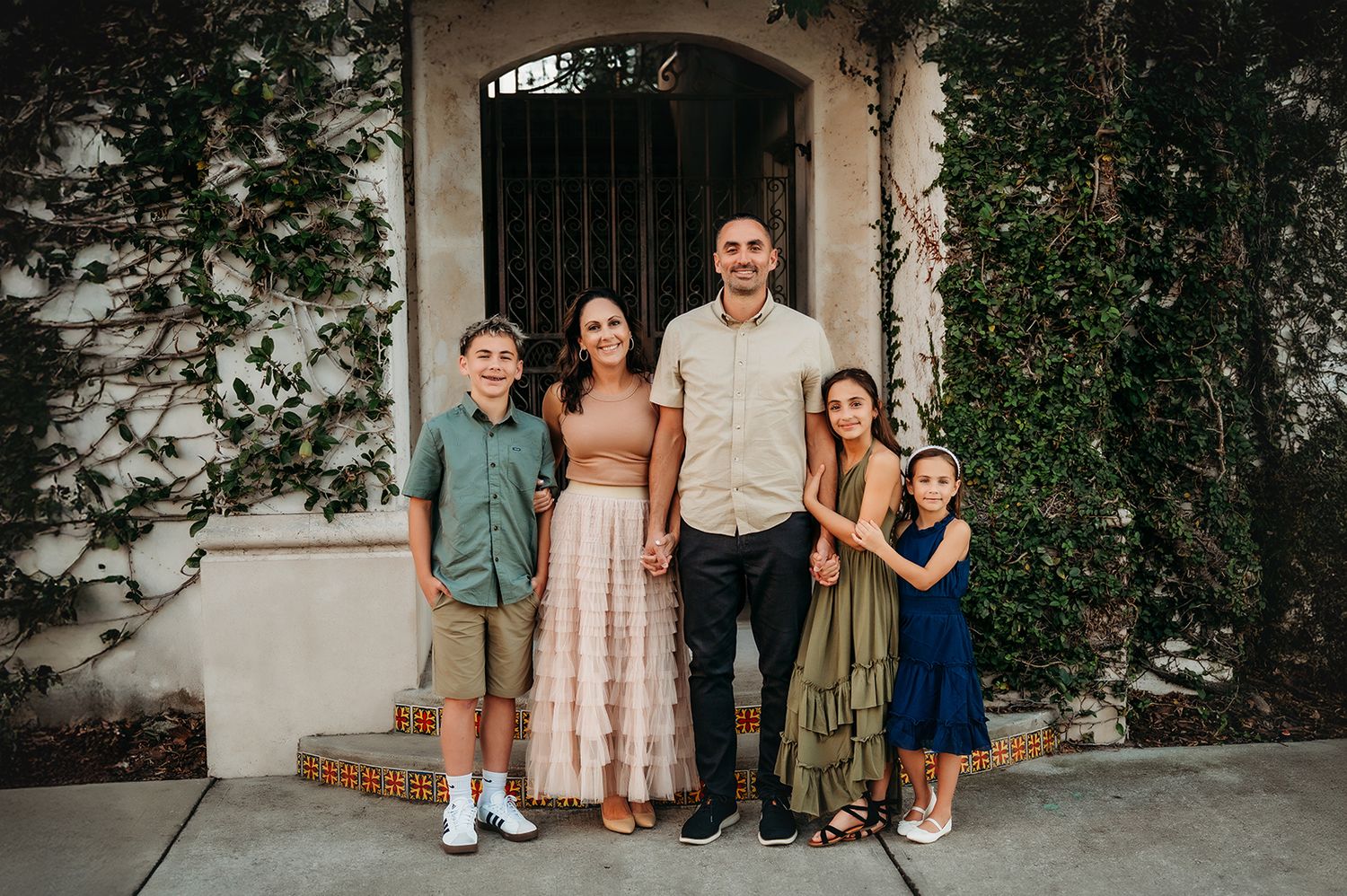 Family of five posing together outdoors in front of an ivy-covered white stucco building.