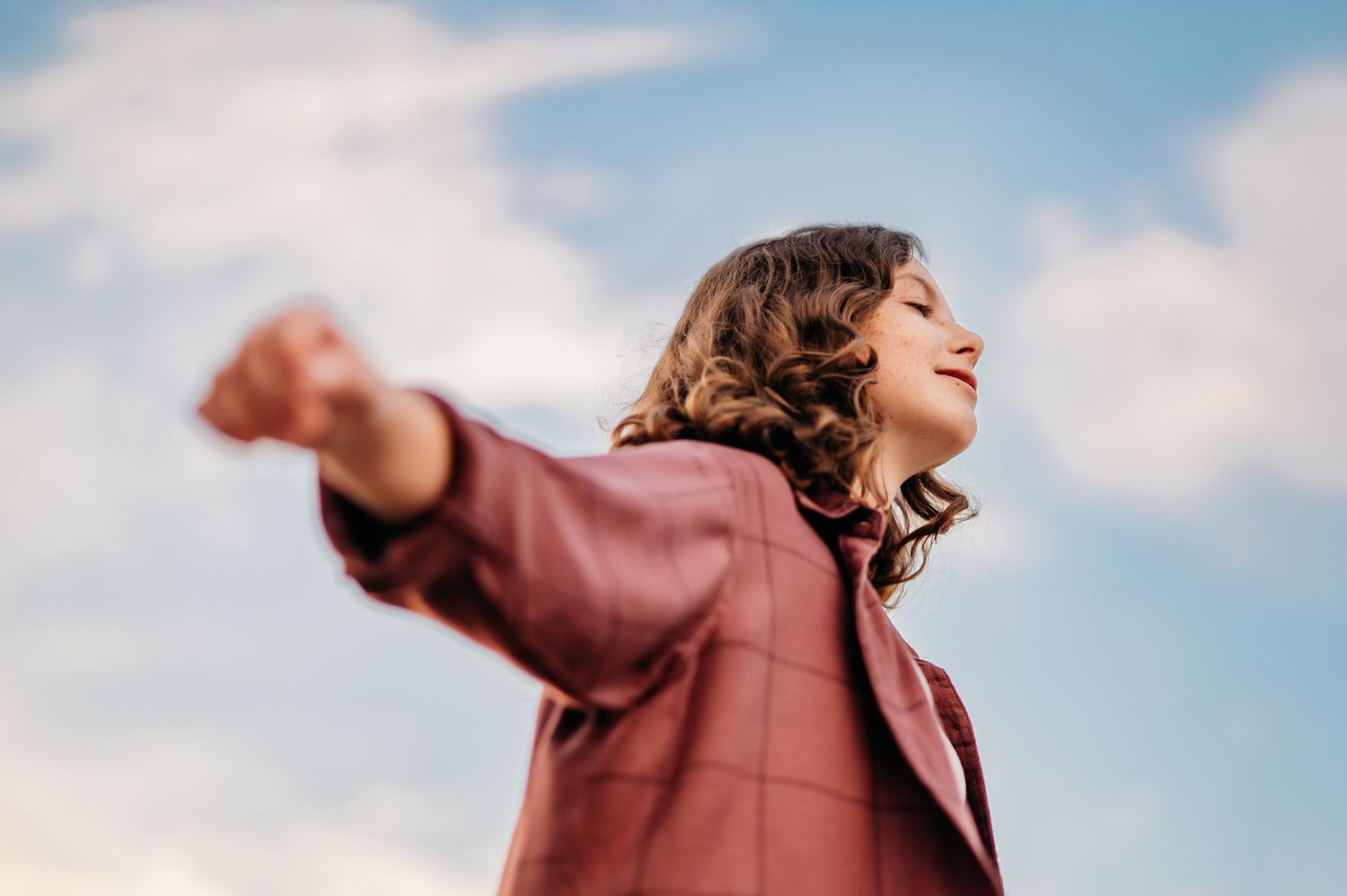 Young woman in a burgundy jacket stretching her arms wide open under a blue sky.