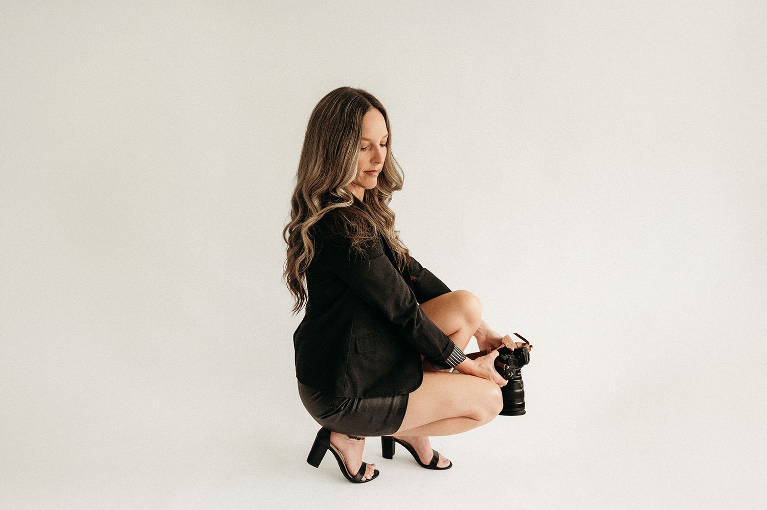 Woman in black dress and heels crouching while holding a professional camera against a white background.