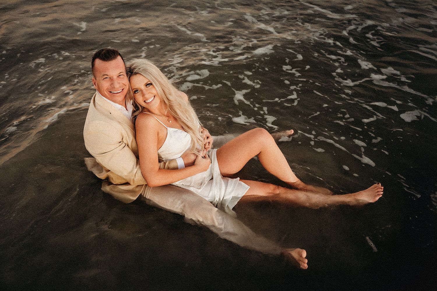 Couple sitting together in shallow ocean waves on a beach, woman in white dress leaning against man in beige shirt.
