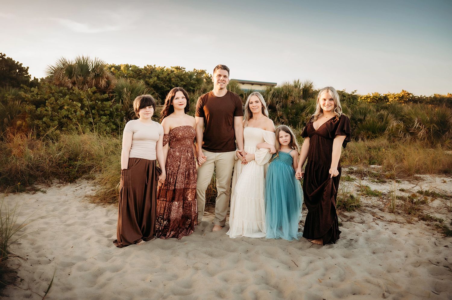 Family of six posing together on sandy beach at golden hour, wearing coordinated earth tones and blue dress.