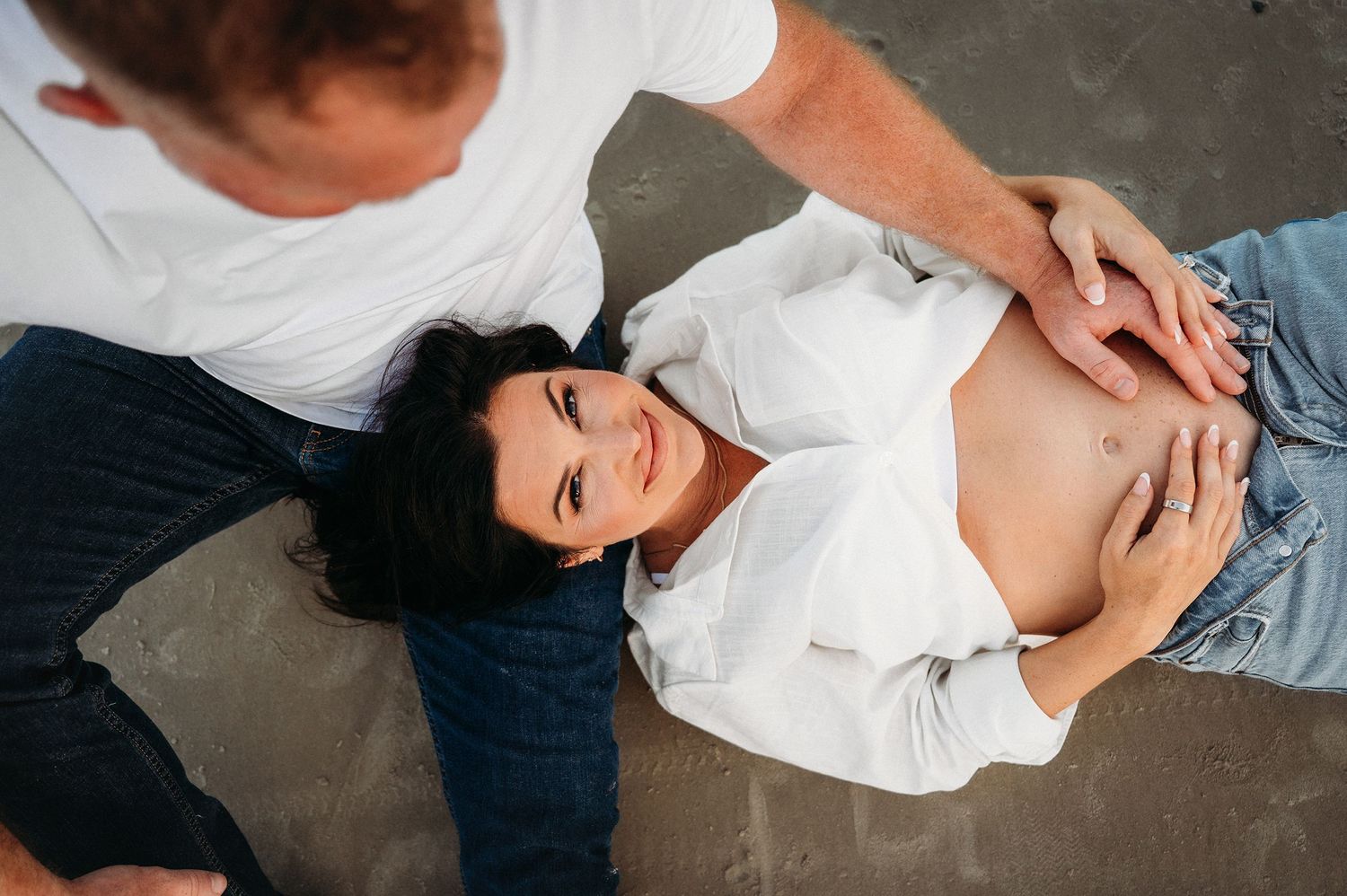 Pregnant woman lying down outdoors while partner tenderly touches her bare baby bump during maternity photoshoot.