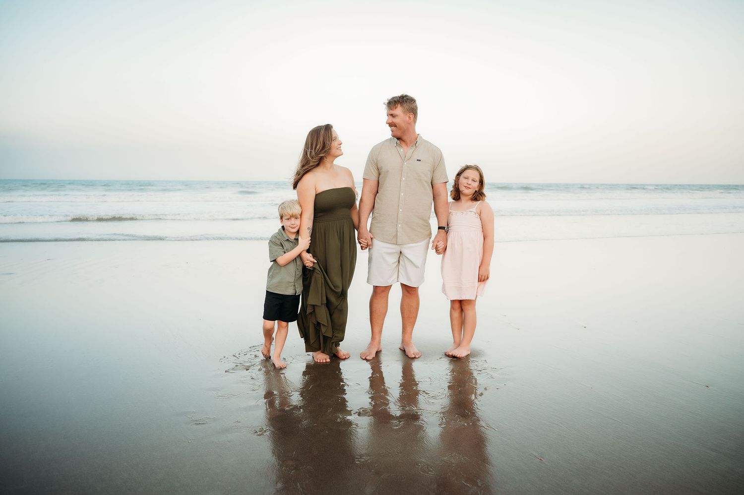 Family of four walking barefoot on a beach at sunset, smiling and holding hands near the ocean shore.