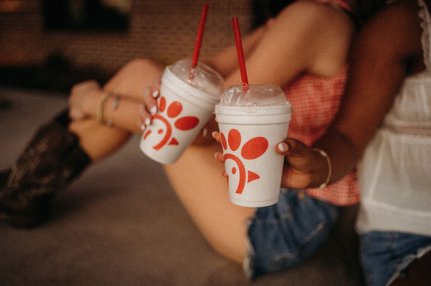 Two people clink red-straw plastic cups with red logos together while sitting close, one wearing cowboy boots and denim shorts.
