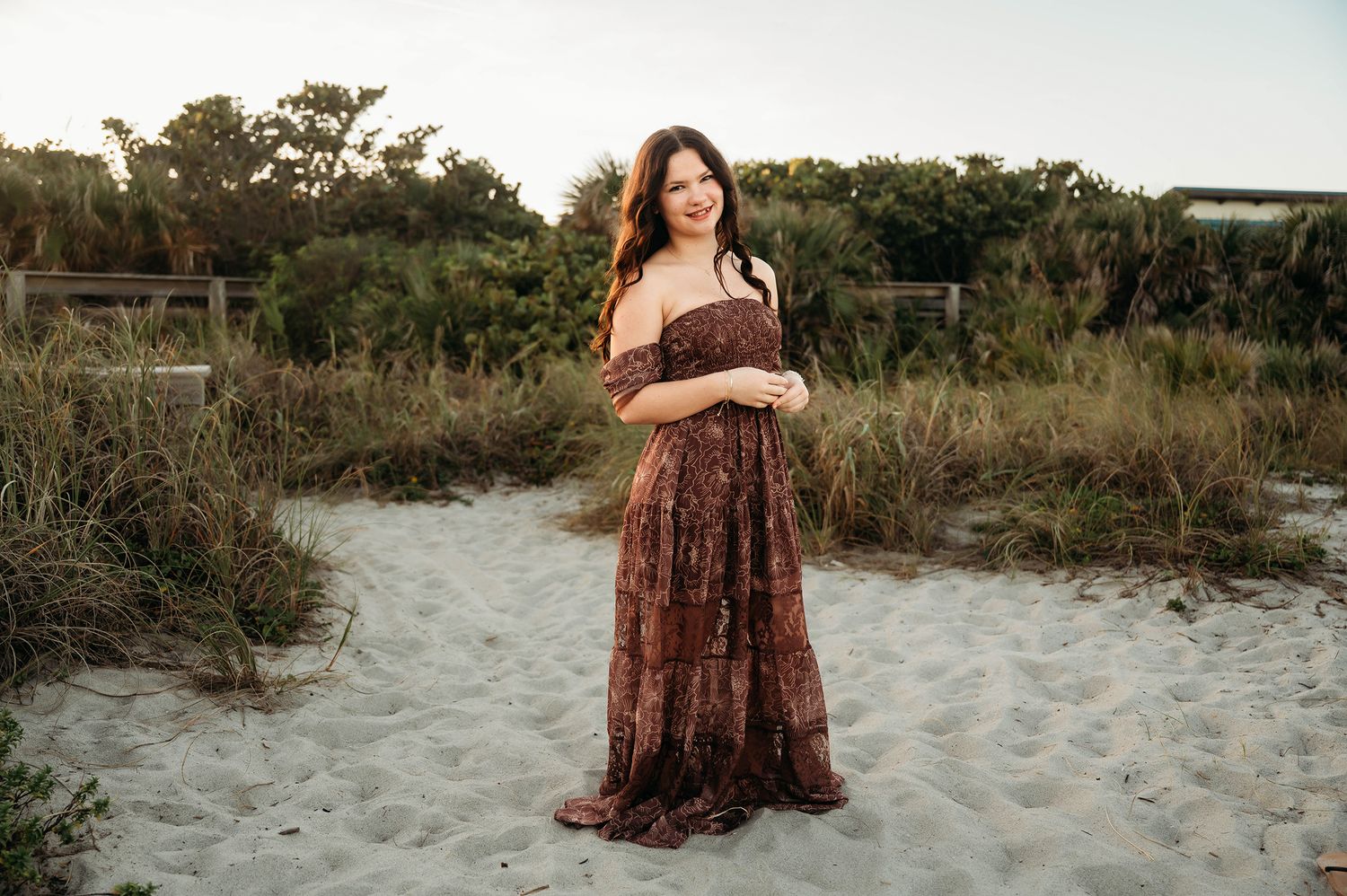A woman in a floral off-shoulder maxi dress smiles on a sandy beach path surrounded by lush greenery.