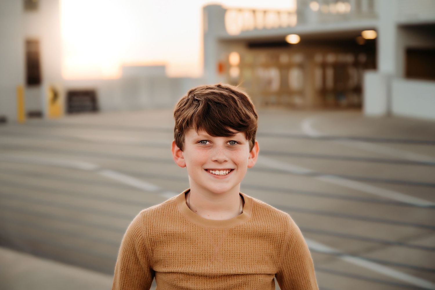 Smiling young boy in a tan ribbed sweater poses on a rooftop parking structure at sunset.