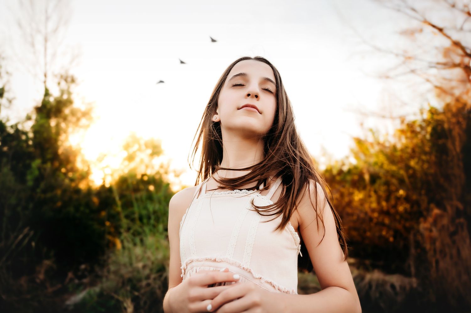 A young girl with long brown hair gazes upward outdoors at golden sunset, birds flying above in warm autumn light.