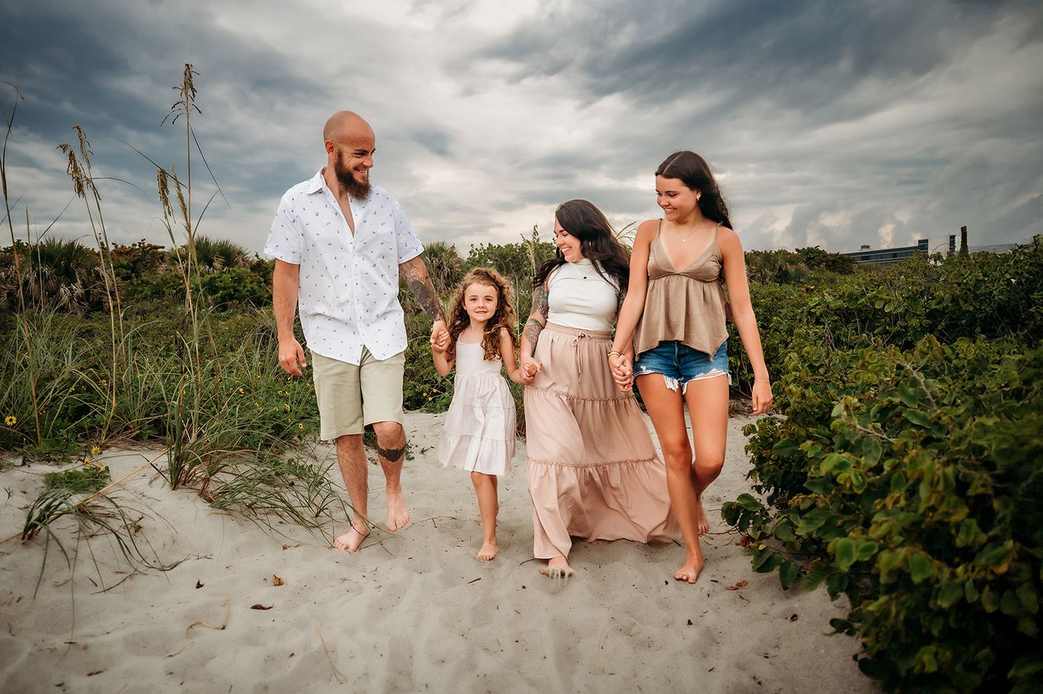 Family of four walking barefoot on cocoa beach path with lush greenery and cloudy sky backdrop.