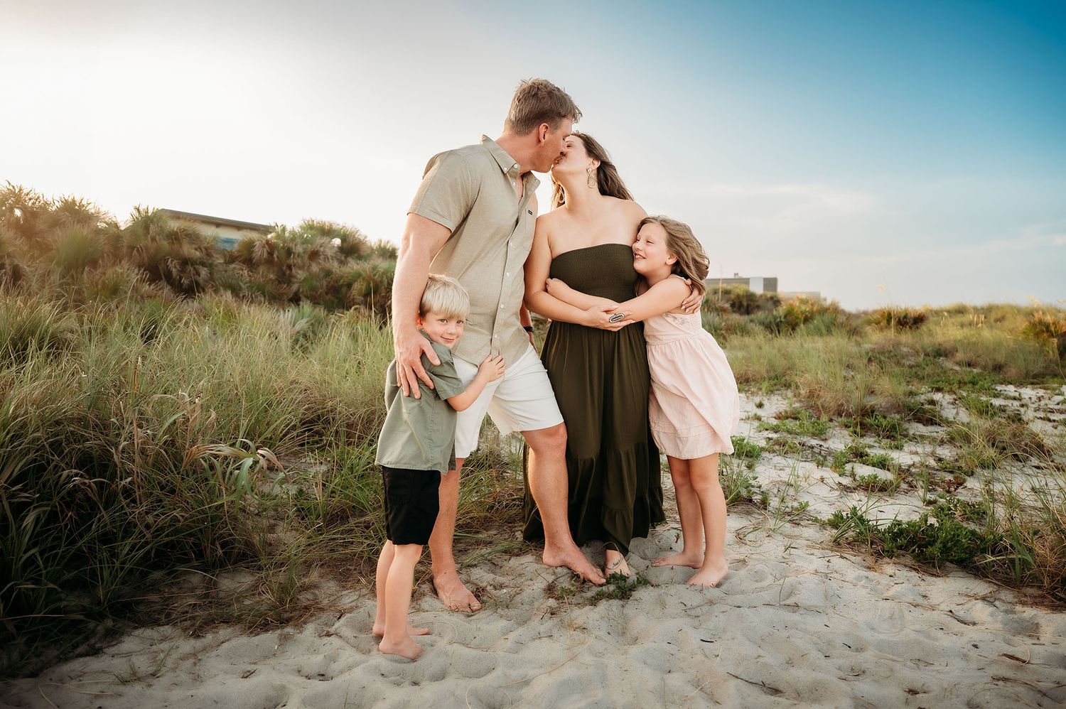 Family of four on sandy beach at sunset, pregnant mother in green dress embraced by husband and two children.