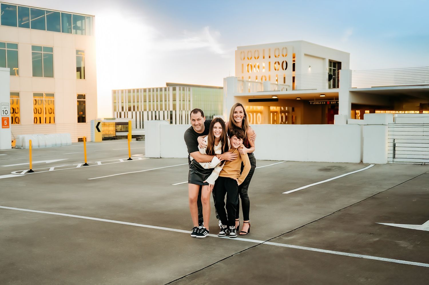Happy family of four posing together on rooftop parking lot at golden hour with modern building behind.
