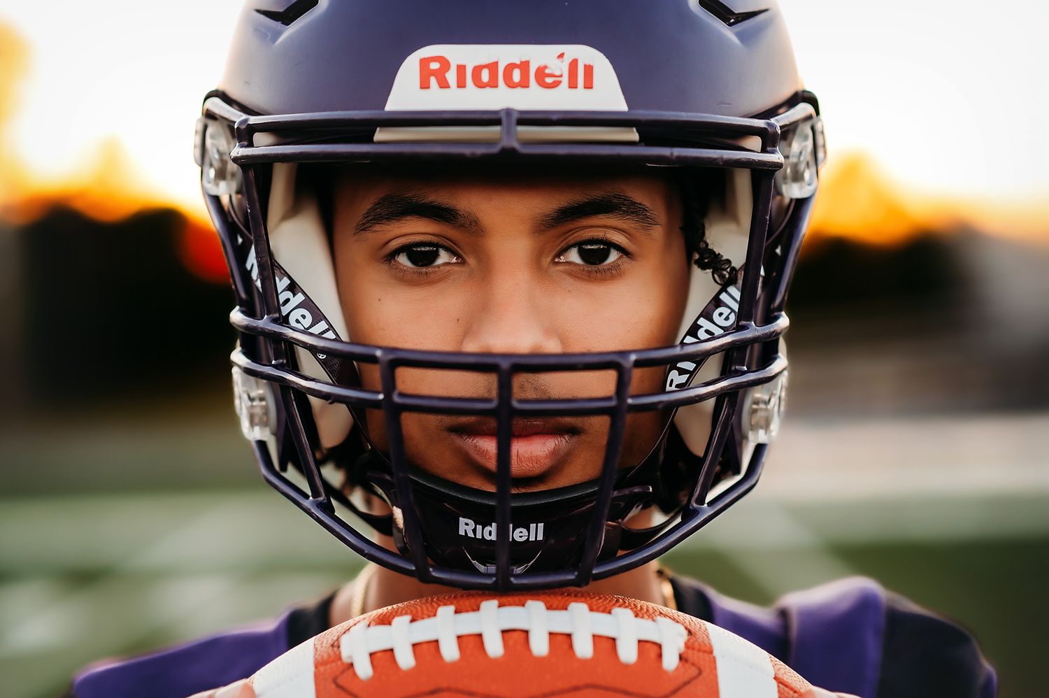 Young football player in purple Riddell helmet holds football, close-up portrait with sunset background.