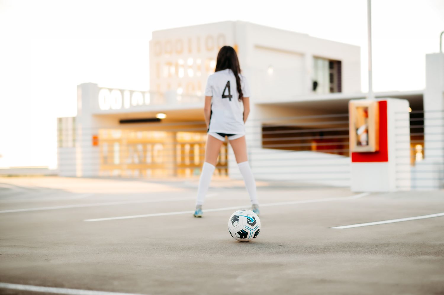 A soccer player in white jersey number 4 and knee-high socks stands behind a ball on an outdoor court.