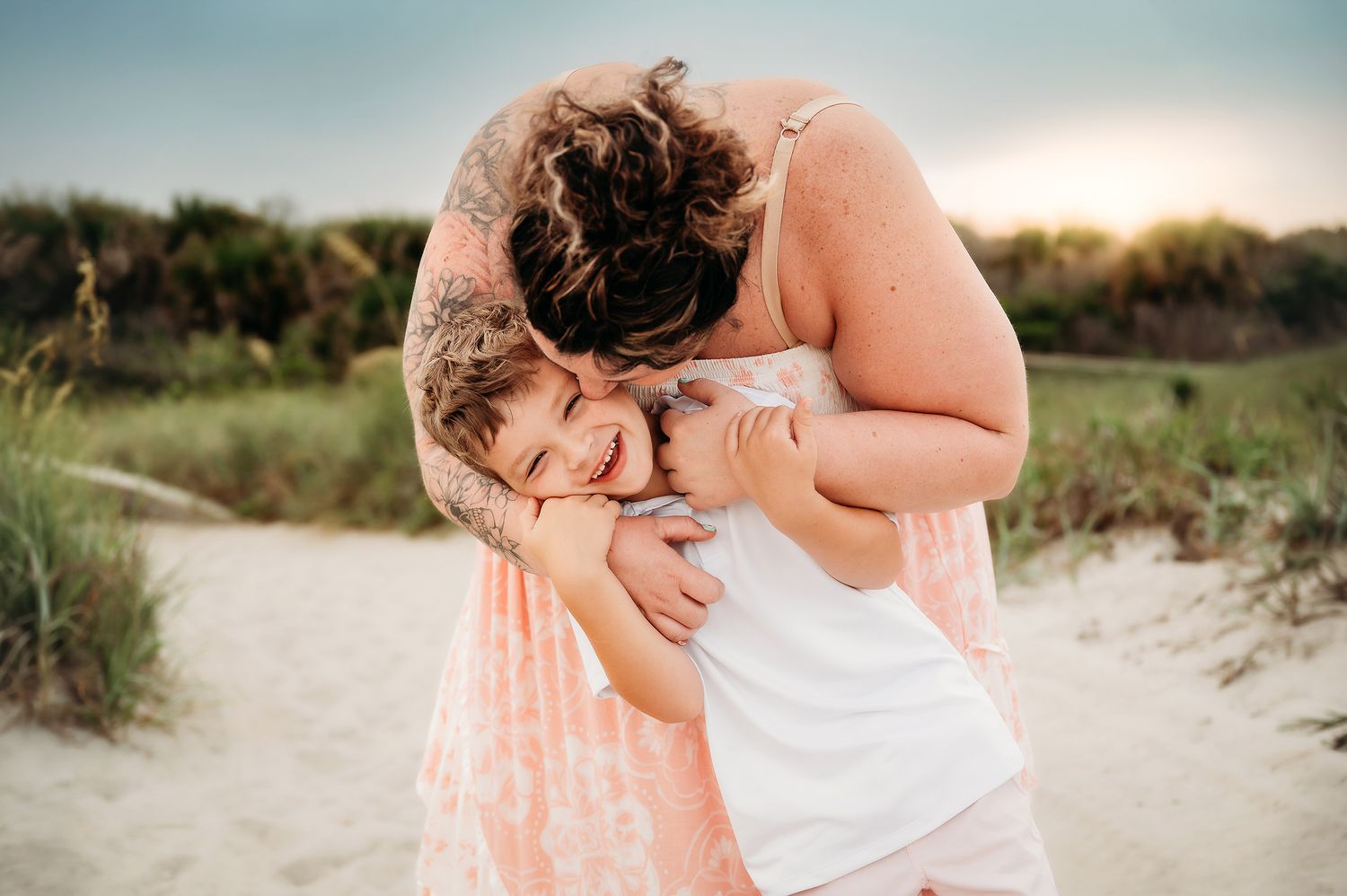 A mother hugging and laughing with her young child on a sandy beach at sunset.