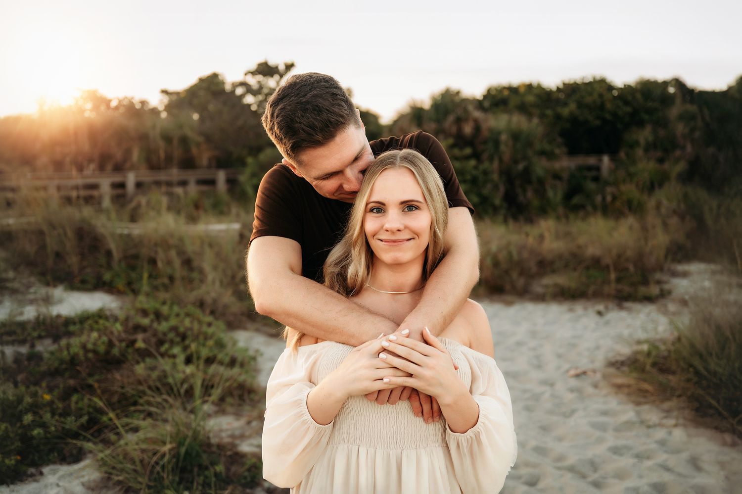 Couple embracing at golden hour on sandy beach, man hugging woman from behind, both smiling warmly with greenery backdrop.