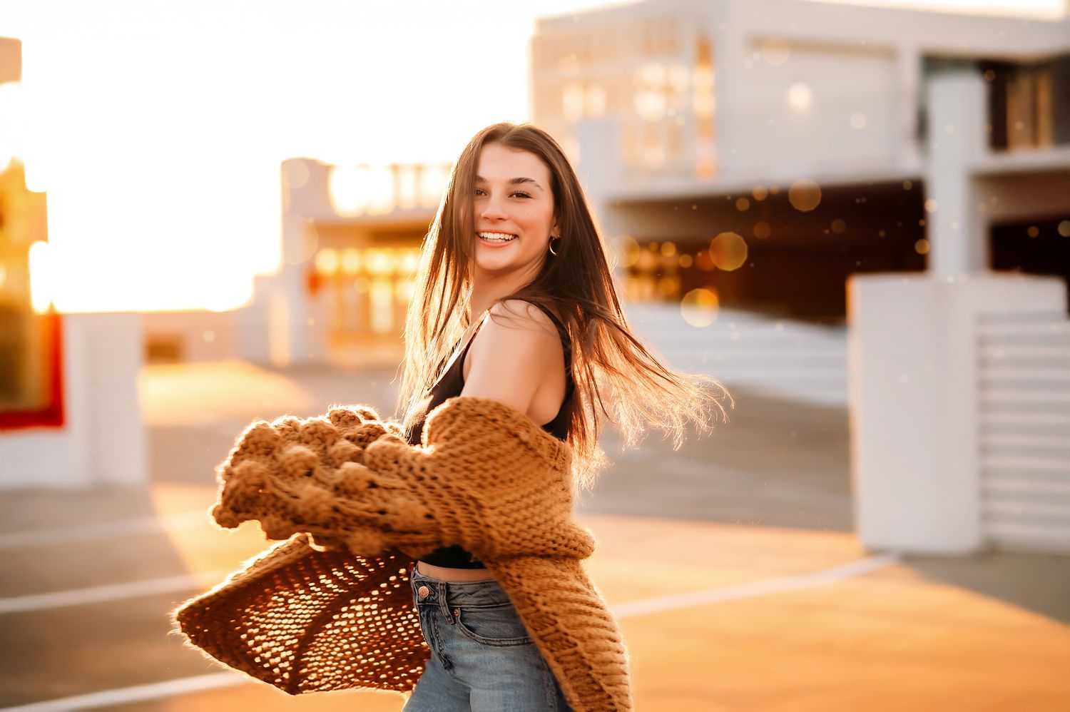 Smiling young woman in jeans and brown knit cardigan holding a woven bag, posing outdoors at golden hour sunset.