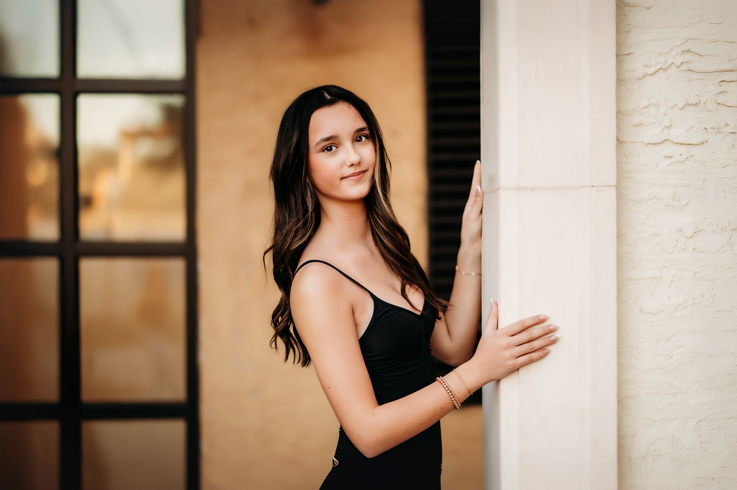 Young woman in black dress posing elegantly against a white column with a warm blurred background.