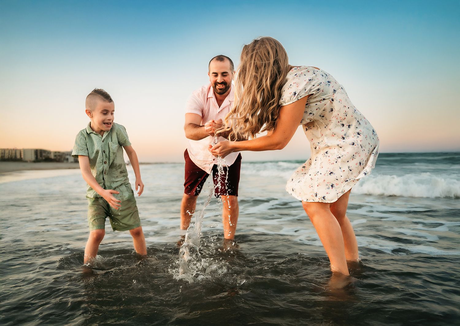 Family of three splashing in ocean waves at sunset on a sandy beach, parents playing with young boy.