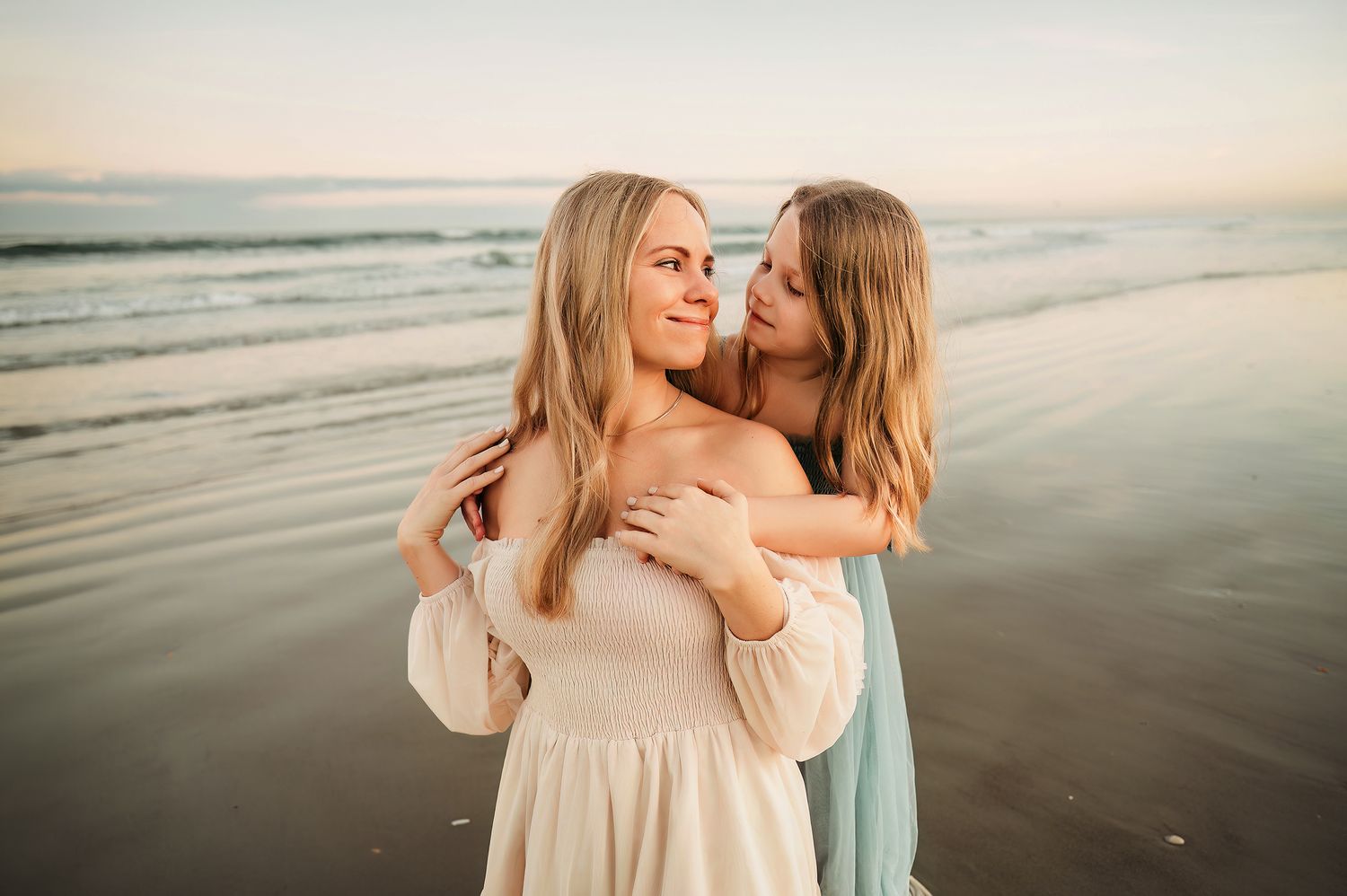Two blonde women embrace on a sandy beach at sunset, wearing flowing dresses with ocean waves in the background.