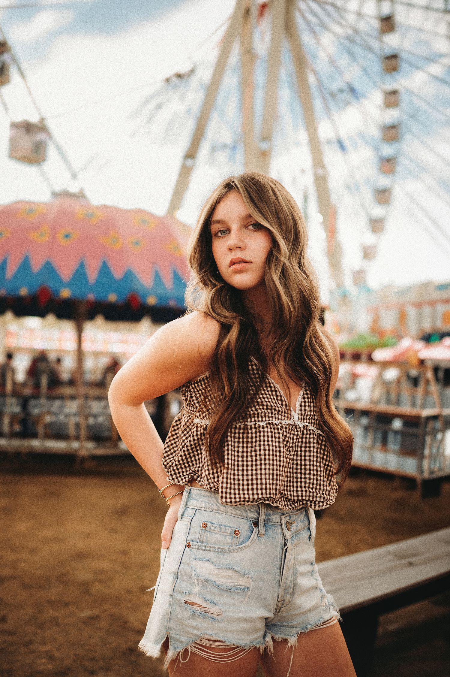 Young woman in checkered top and denim shorts posing at a county fair with a Ferris wheel backdrop.