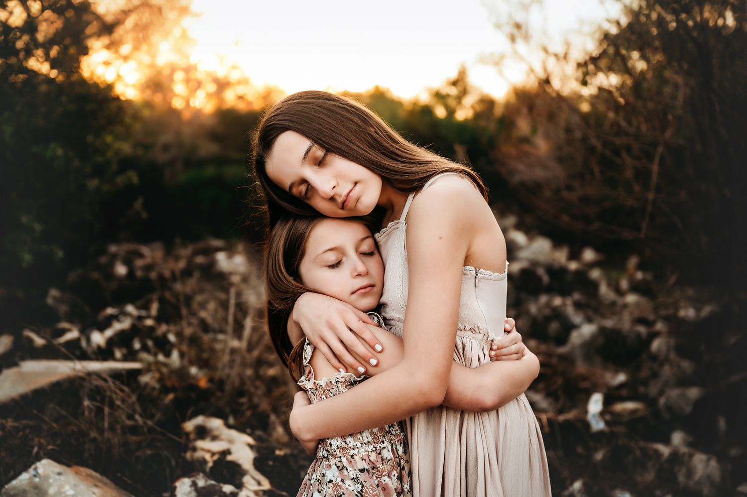 A mother and daughter share a tender embrace outdoors at golden hour, surrounded by nature.