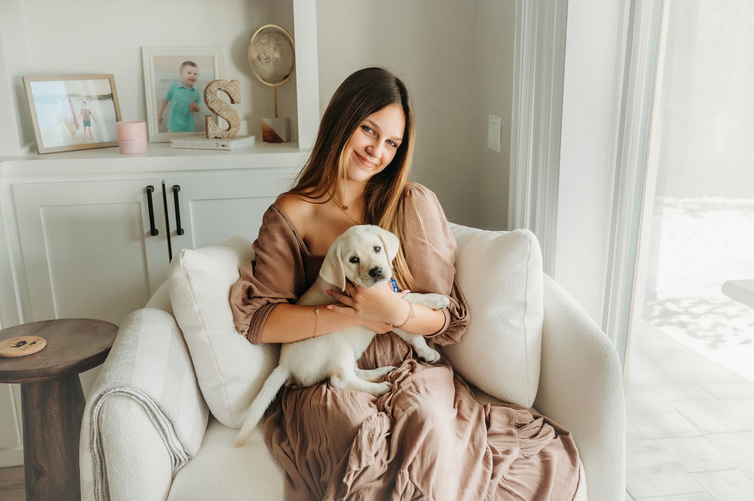 A young woman smiles while holding a white puppy on a cozy white armchair in a bright, elegant living room.