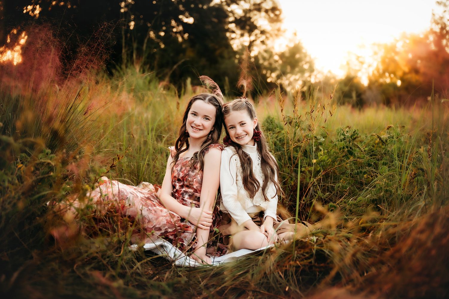 Two smiling girls sit back-to-back in a golden meadow at sunset, wearing floral and white dresses.