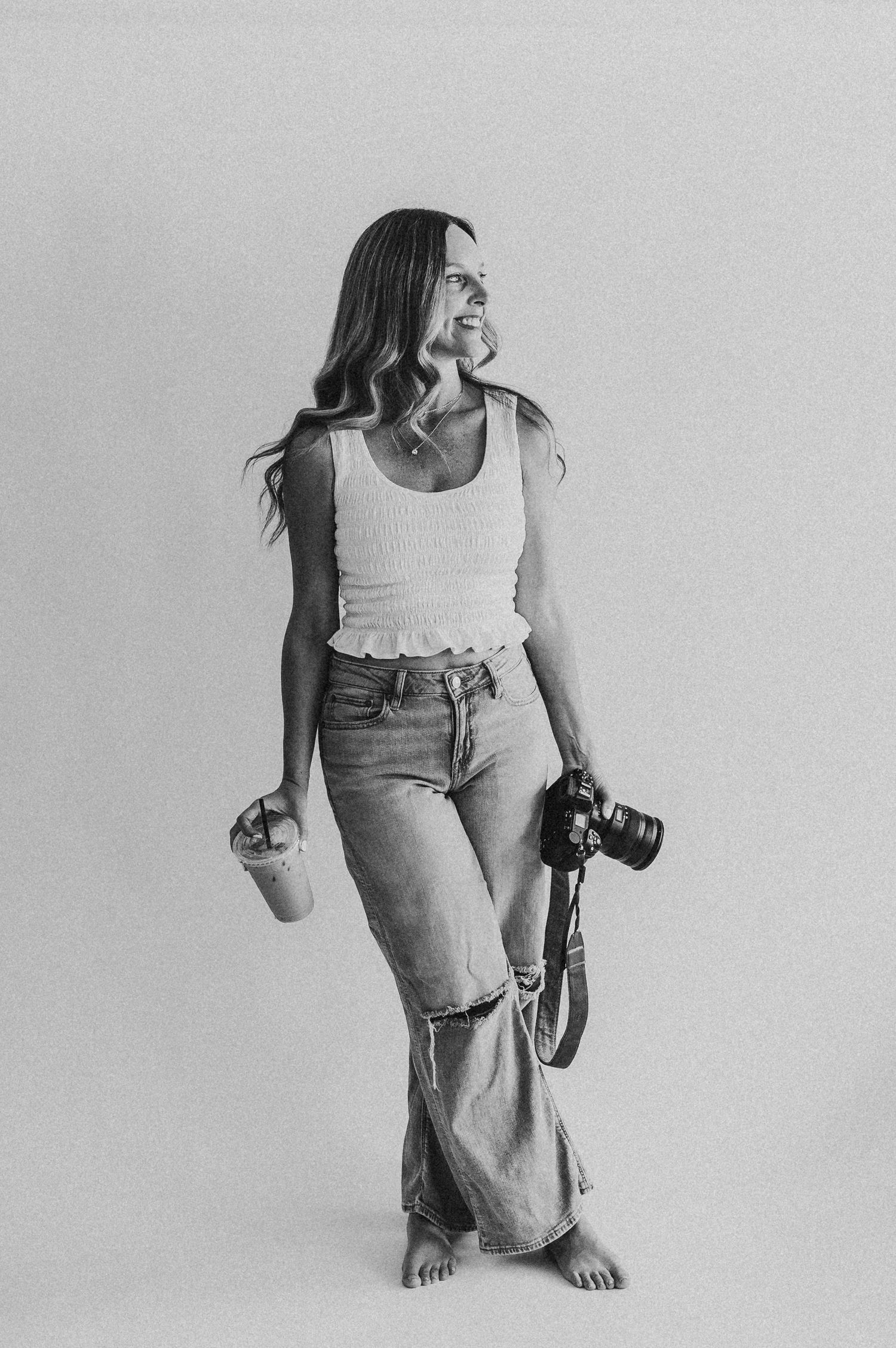Black and white photo of a woman holding a camera and drink, wearing jeans and a white tank top.