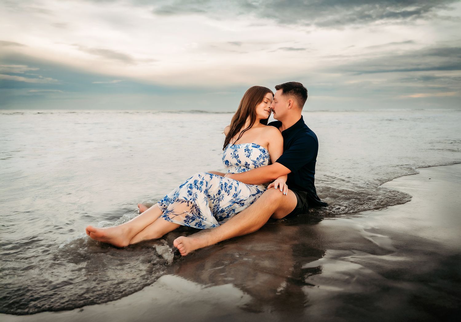 A couple shares a romantic moment sitting together on a wet beach shore at sunset, woman in floral dress.